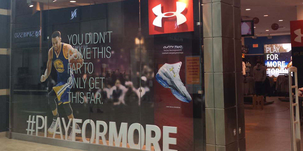 Under Armour store window display featuring Stephen Curry, Curry Two shoes, and the #PLAYFORMORE slogan.