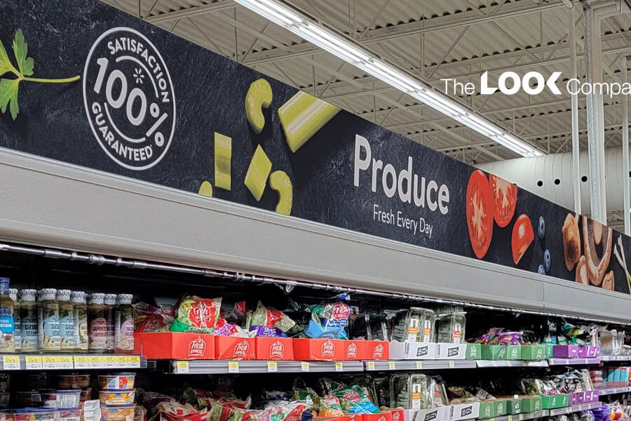A grocery store produce aisle features a long black banner displaying ""Produce Fresh Every Day,"" a ""100% Satisfaction Guaranteed"" logo, and various fruit and vegetable images.
