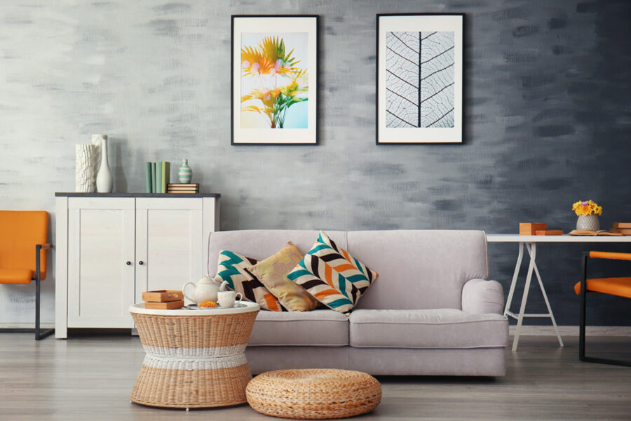 A modern living room features a light grey sofa with patterned pillows, a wicker coffee table, orange accent chairs, and nature-themed framed art on a textured grey wall.