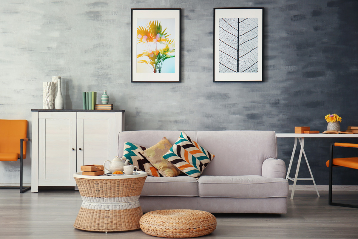 A modern living room features a light grey sofa with patterned pillows, a wicker coffee table, orange accent chairs, and nature-themed framed art on a textured grey wall.