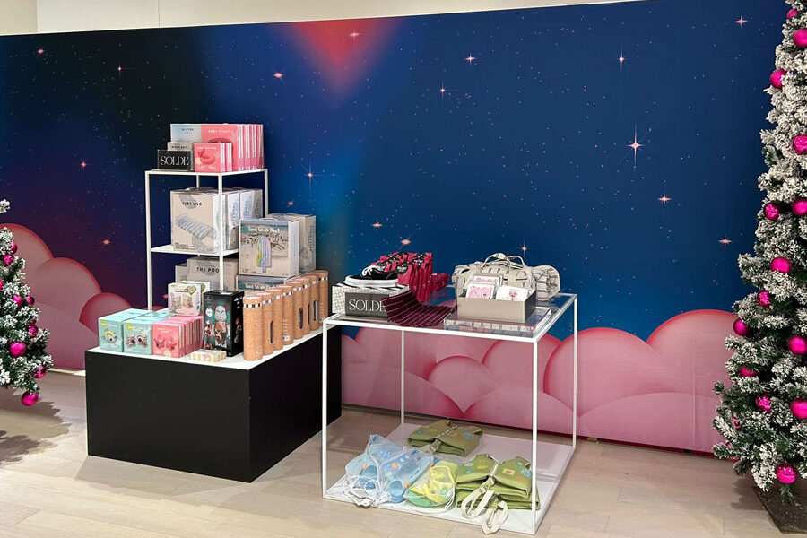 A festive retail display features products on shelves and tables between two snow-dusted Christmas trees with pink ornaments, set against a starry blue backdrop with pink clouds.