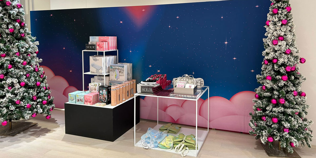 A festive retail display features products on shelves and tables between two snow-dusted Christmas trees with pink ornaments, set against a starry blue backdrop with pink clouds.
