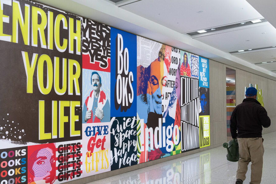 A long, vibrant mural composed of typographic art and portraits, including 'ENRICH YOUR LIFE' and 'BOOKS,' lines a bright public hallway with people passing.