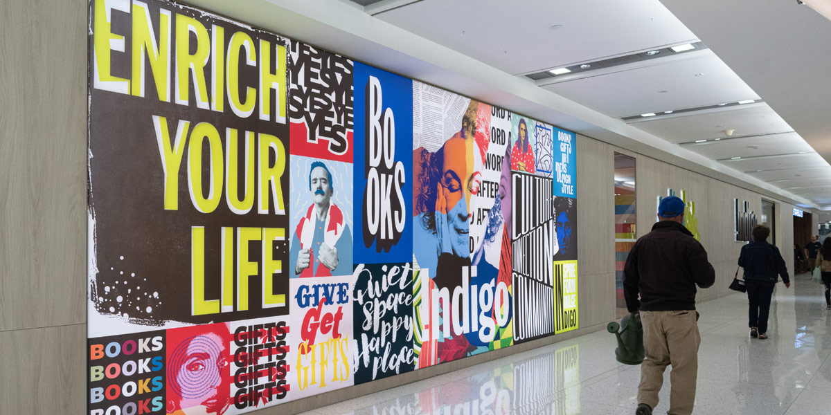 A long, vibrant mural composed of typographic art and portraits, including 'ENRICH YOUR LIFE' and 'BOOKS,' lines a bright public hallway with people passing.