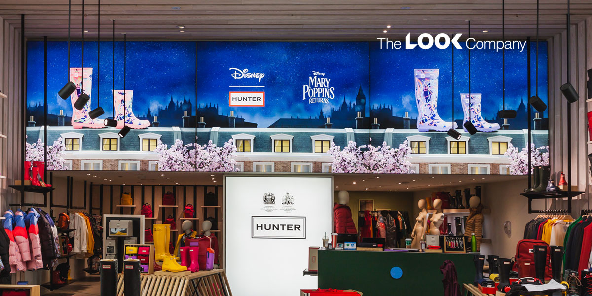 A vibrant Hunter retail store interior features a large digital display promoting a Disney Mary Poppins Returns collaboration with stylized rain boots, above colorful clothing and footwear merchandise.