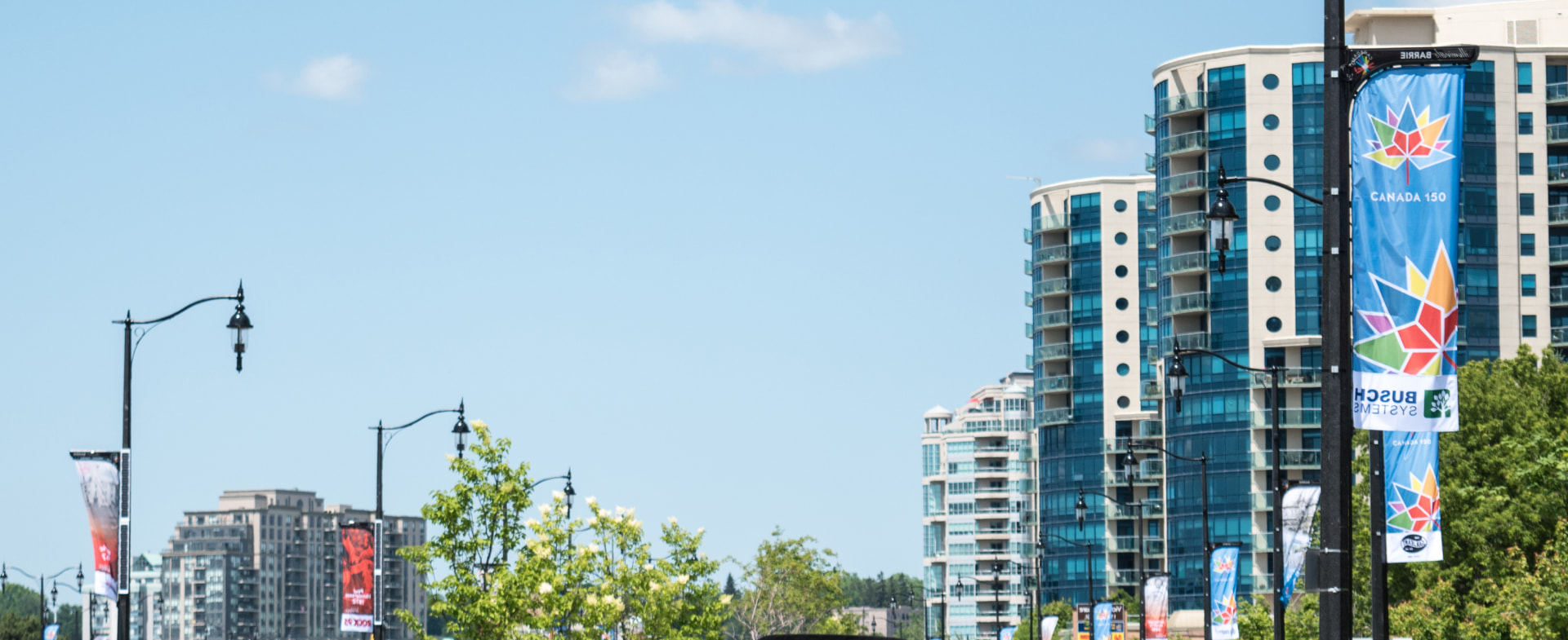Modern high-rise buildings line a street adorned with blue ""Canada 150"" banners under a clear sky.