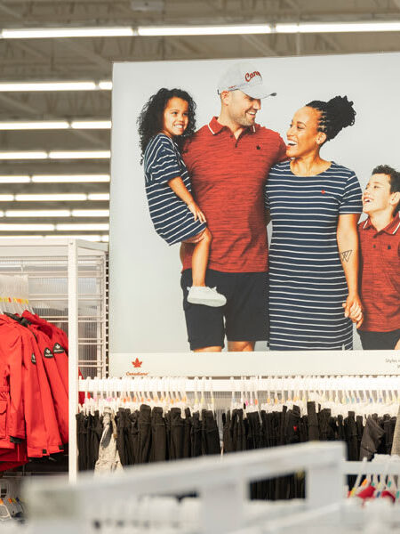 "A large advertisement poster featuring a smiling diverse family in matching red and striped clothing hangs in a retail store's apparel section, surrounded by racks of clothing."