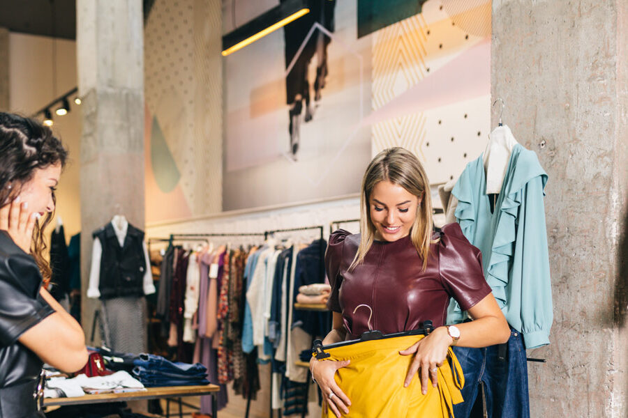 Two women are clothes shopping in a store; one holds up a yellow skirt and smiles as the other looks on.