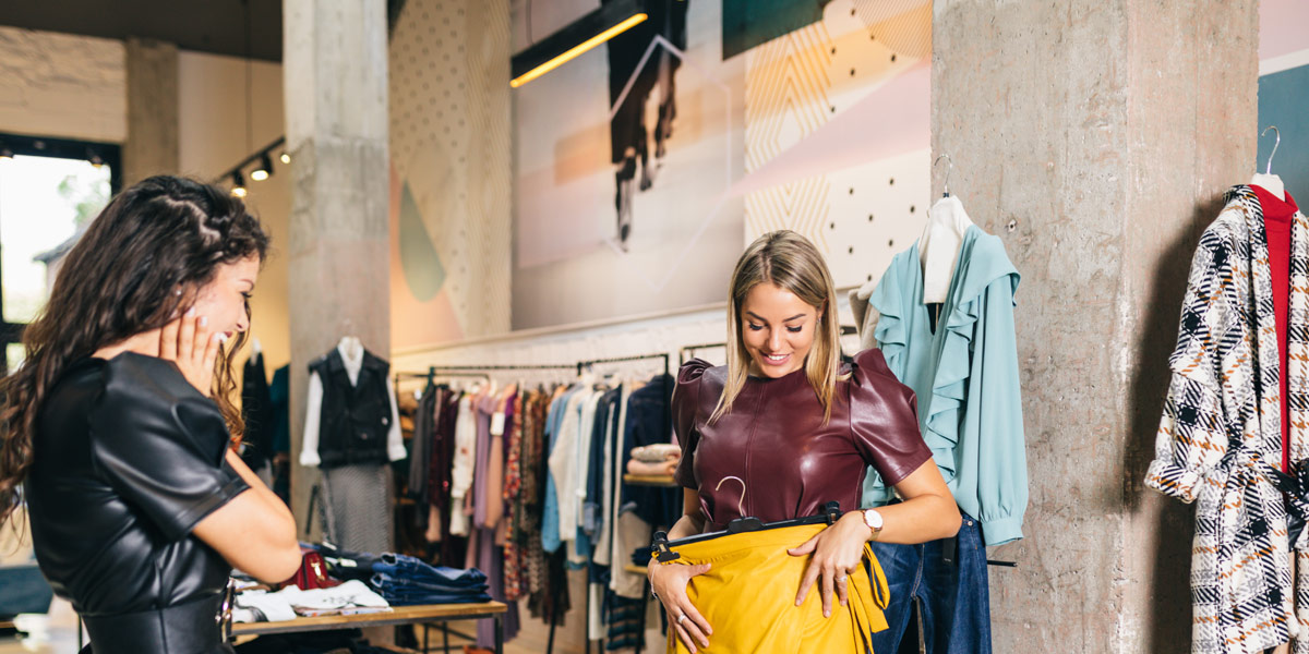 Two women are clothes shopping in a store; one holds up a yellow skirt and smiles as the other looks on.