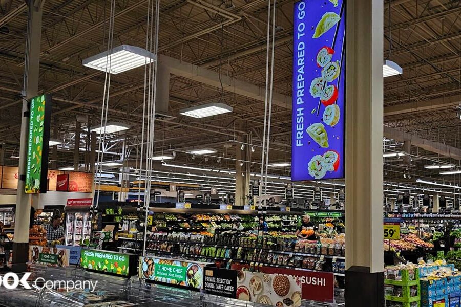 Interior of a brightly lit supermarket featuring a ""Fresh Prepared To Go"" section with hanging digital signs, fresh produce, and packaged meals.