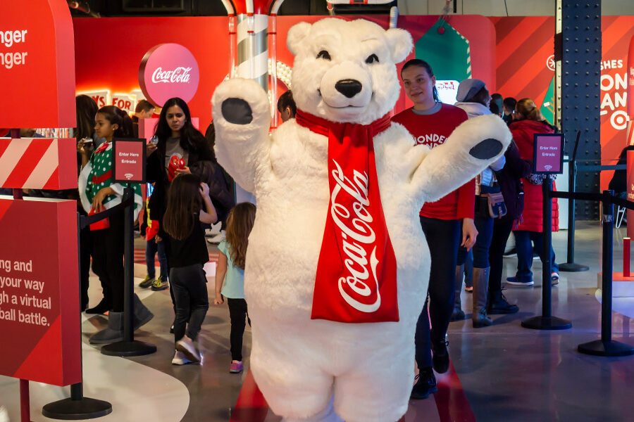 A Coca-Cola polar bear mascot waves to guests in a festive red-themed event space advertising a ""Slinger Flinger"" virtual snowball battle.