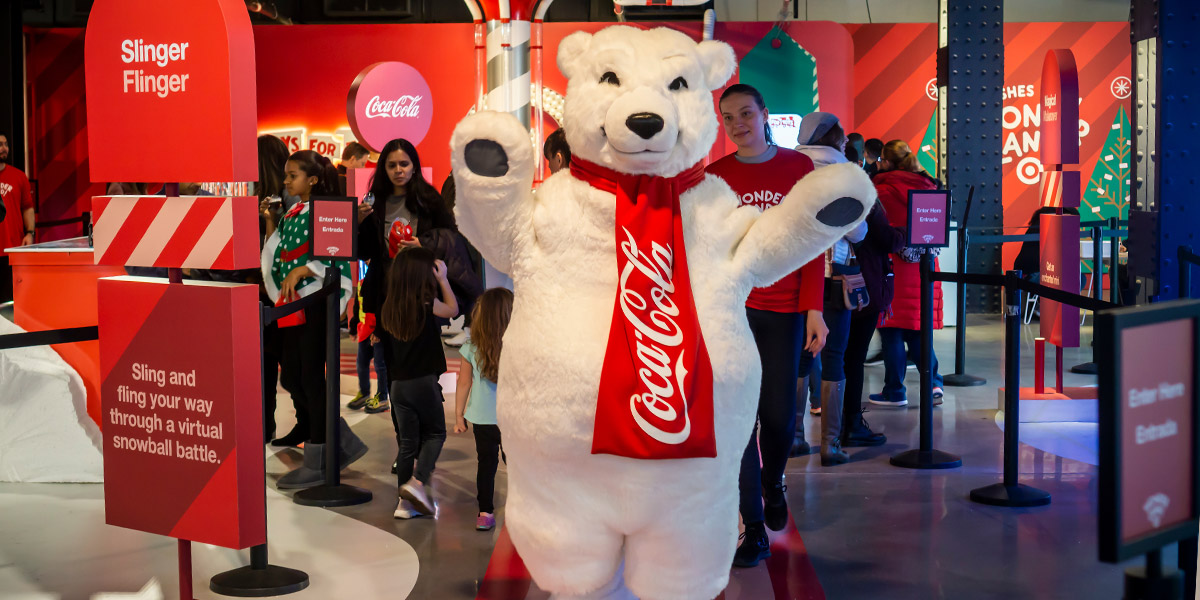 A Coca-Cola polar bear mascot waves to guests in a festive red-themed event space advertising a ""Slinger Flinger"" virtual snowball battle.