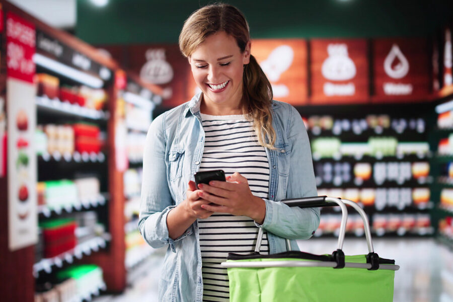 A smiling woman looks at her smartphone while holding a green shopping basket in a grocery store aisle.