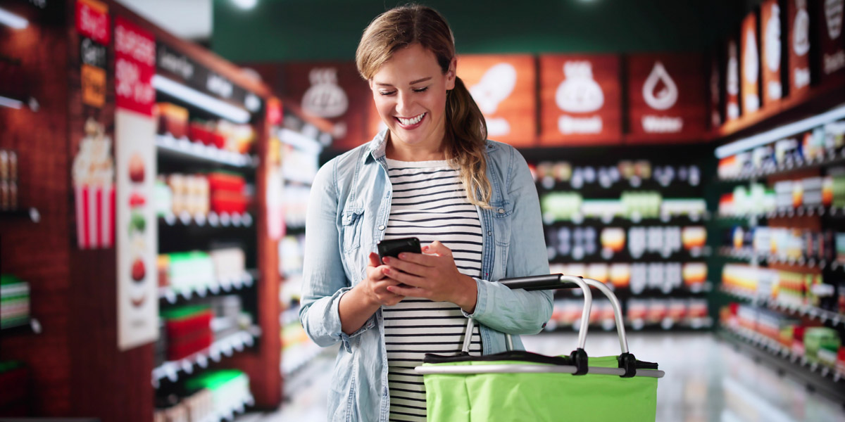A smiling woman looks at her smartphone while holding a green shopping basket in a grocery store aisle.
