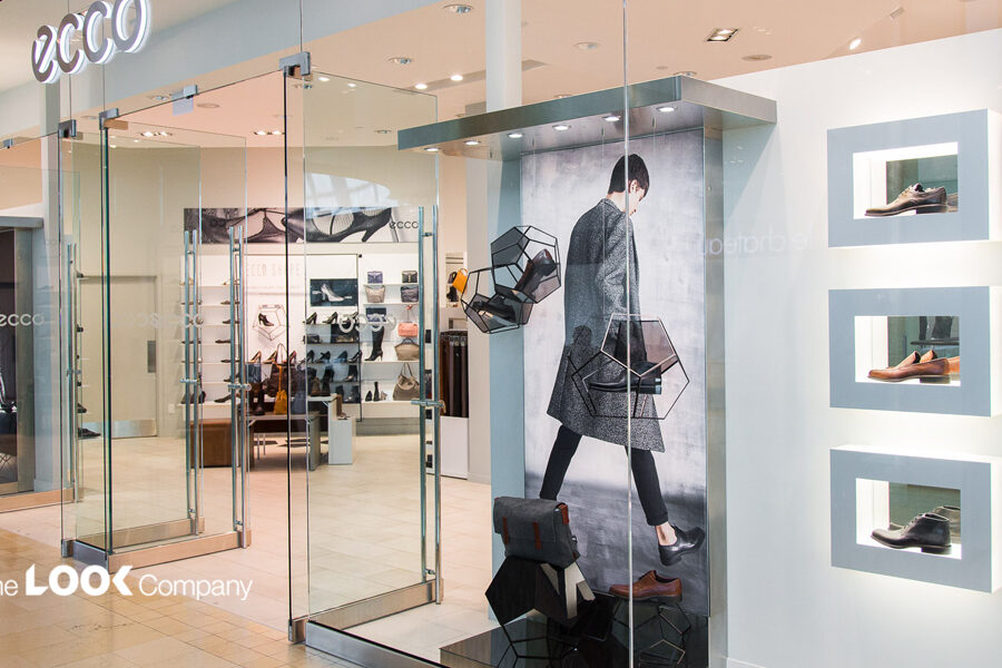 The bright, modern Ecco shoe store features glass walls displaying shoes in geometric frames and illuminated wall recesses, with additional footwear visible inside.