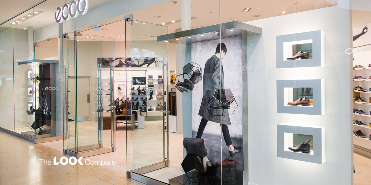 The bright, modern Ecco shoe store features glass walls displaying shoes in geometric frames and illuminated wall recesses, with additional footwear visible inside.