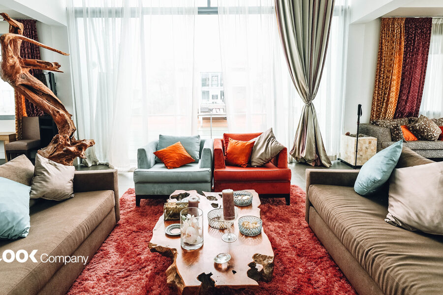 A brightly lit, modern living room features a large driftwood sculpture, colorful sofas, a live-edge coffee table, and a vibrant red shag rug.