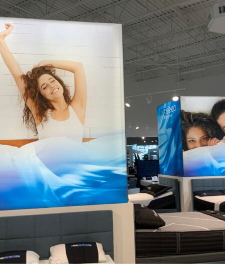 A bright mattress showroom features large promotional displays of people in bed, surrounded by numerous bed frames and mattresses.