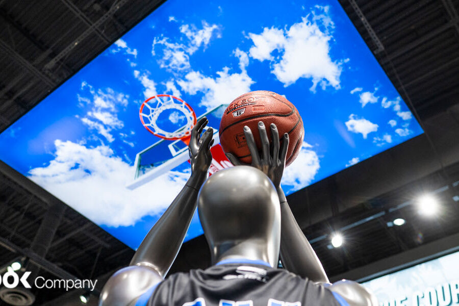 A grey mannequin holds a basketball, poised to shoot at a hoop displayed on a large ceiling screen featuring a blue sky with clouds.