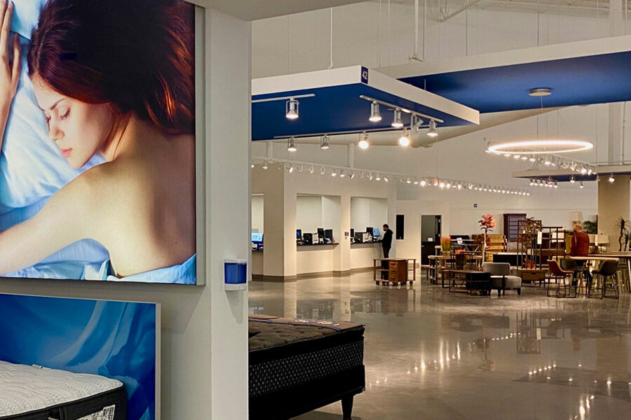 Modern mattress and furniture showroom with a large advertisement of a sleeping woman, product displays, and customers browsing.