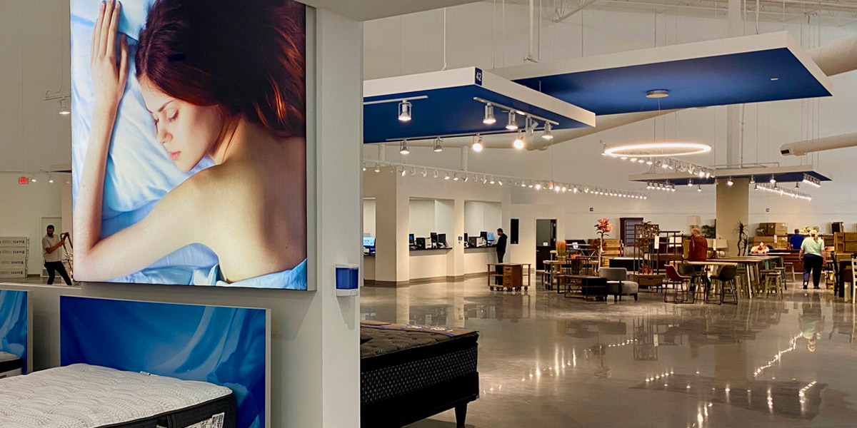 Modern mattress and furniture showroom with a large advertisement of a sleeping woman, product displays, and customers browsing.
