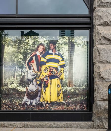 A Louis Vuitton store window displays a vibrant ad featuring two models in colorful, patterned outfits against a forest backdrop, framed by a stone building and a parking meter on a city street.