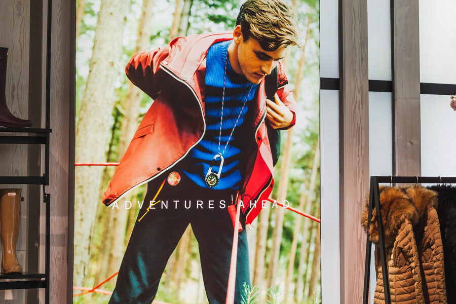 A vibrant retail display features a man in outdoor gear on a large poster, alongside shelves of tall boots, a rack of winter coats, and decorative pink cherry blossoms.