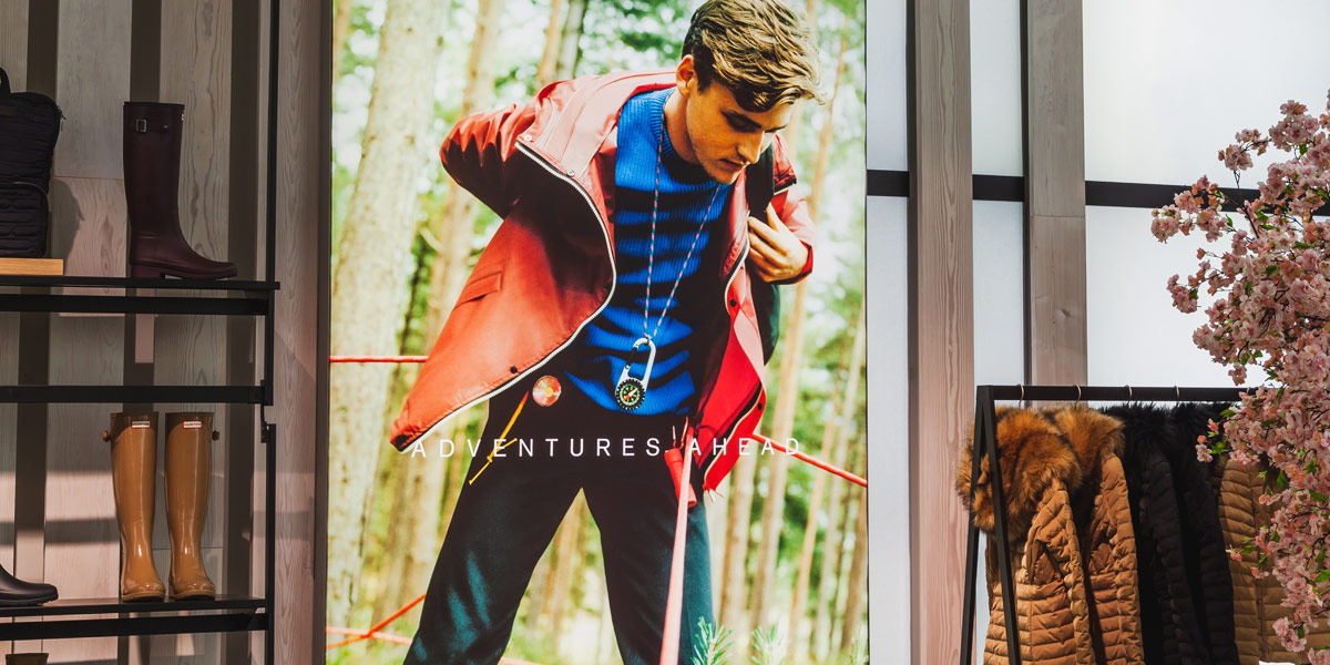 A vibrant retail display features a man in outdoor gear on a large poster, alongside shelves of tall boots, a rack of winter coats, and decorative pink cherry blossoms.