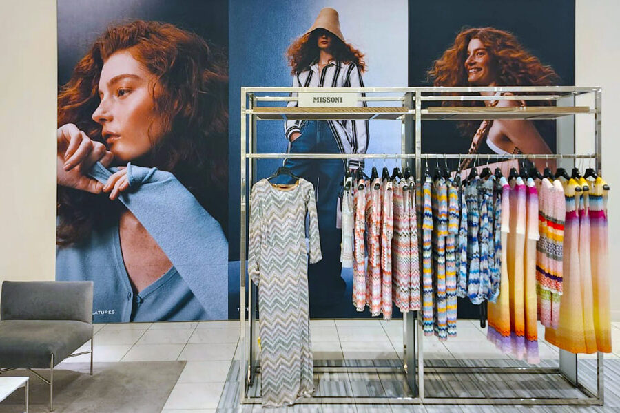 Missoni clothing display featuring colorful patterned knitwear on a rack, set against large fashion model posters.