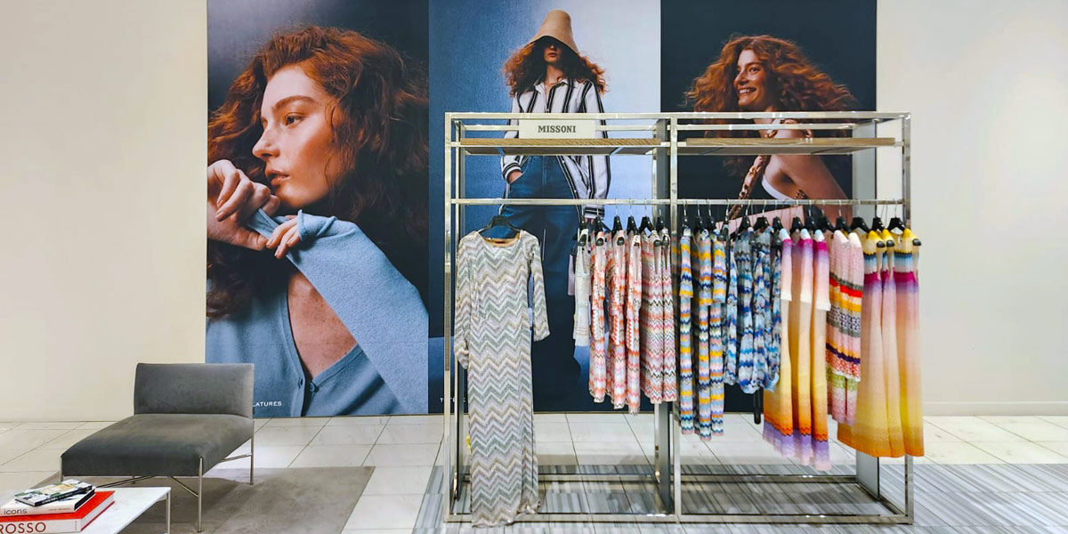Missoni clothing display featuring colorful patterned knitwear on a rack, set against large fashion model posters.