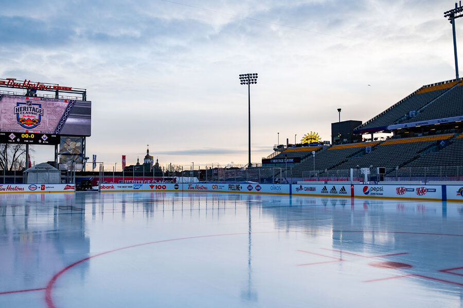 An empty outdoor hockey rink is ready in a stadium, with a scoreboard displaying the ""Heritage Classic"" logo and vacant stands under a cloudy sky.