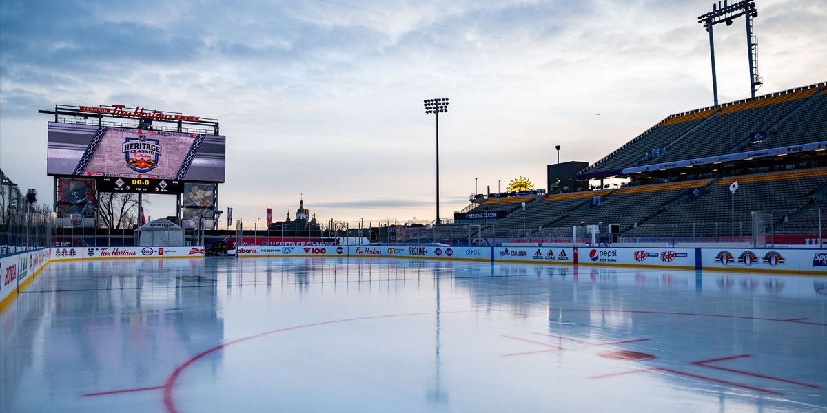 An empty outdoor hockey rink is ready in a stadium, with a scoreboard displaying the ""Heritage Classic"" logo and vacant stands under a cloudy sky.