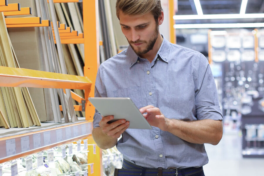A man uses a tablet in a hardware store aisle with shelves stocked with building materials.