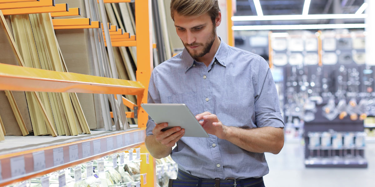 A man uses a tablet in a hardware store aisle with shelves stocked with building materials.