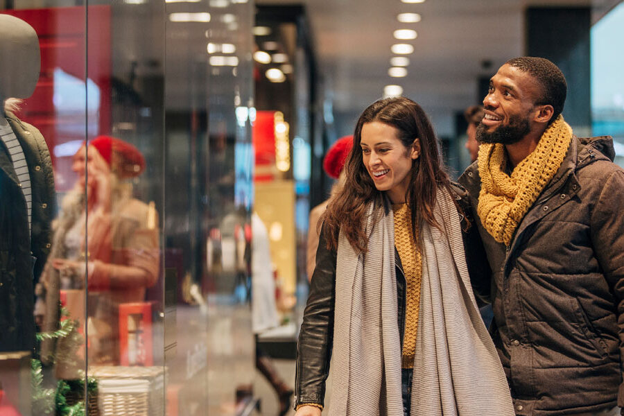A smiling man and woman walk past brightly lit store windows, appearing to window shop.
