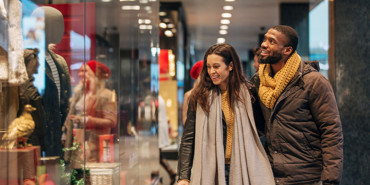 A smiling man and woman walk past brightly lit store windows, appearing to window shop.