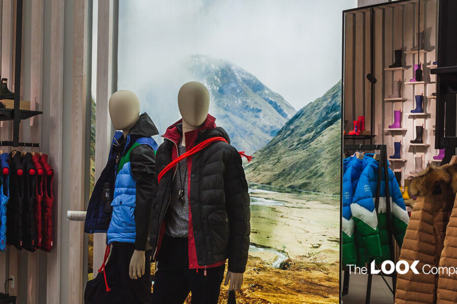 A retail store display featuring mannequins dressed in outdoor jackets, colorful apparel on racks, rubber boots on shelves, and a scenic mountain backdrop, with 'The LOOK Company' logo visible.