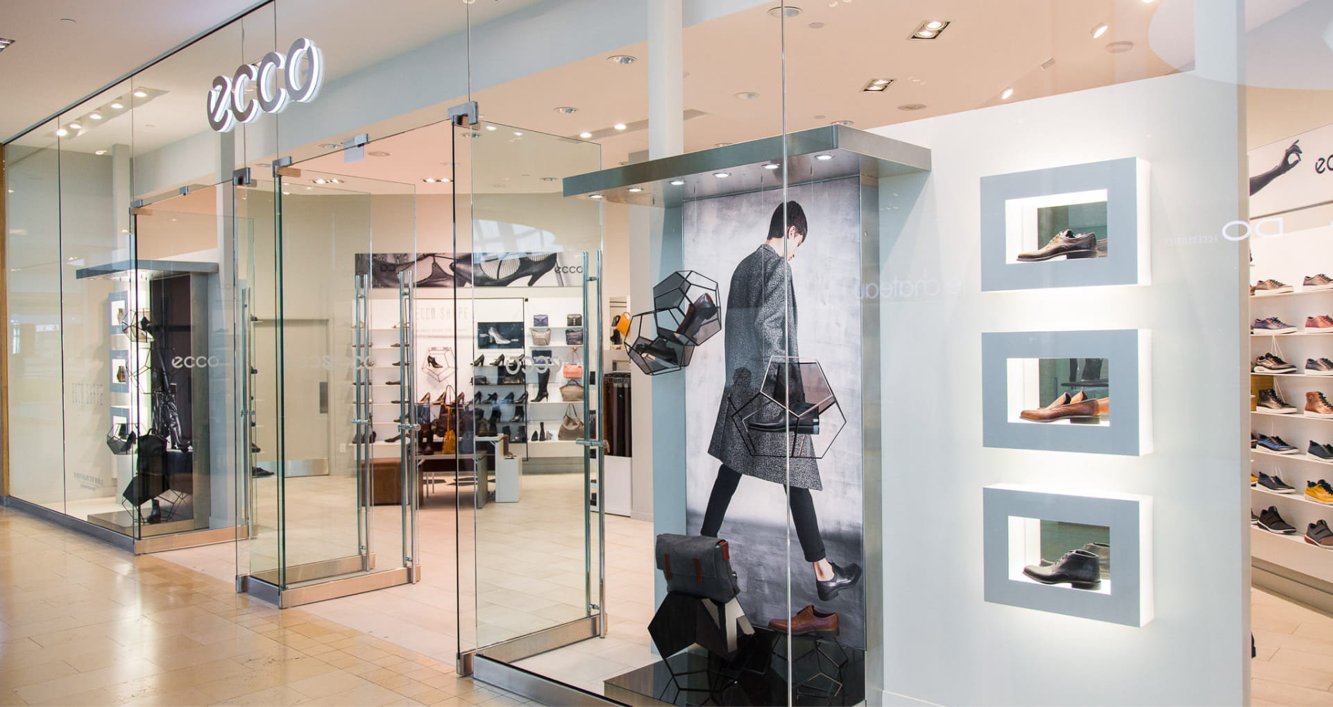 A bright, modern ECCO retail store entrance with floor-to-ceiling glass walls. The interior is well-lit, featuring minimalist displays of shoes and bags. A large window graphic shows a person walking, and the white walls feature recessed shelving highlighting specific footwear styles.