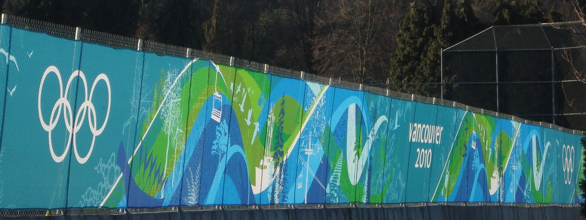 A long blue and green fence banner for the Vancouver 2010 Olympics features the Olympic rings and a stylized mountain landscape design.