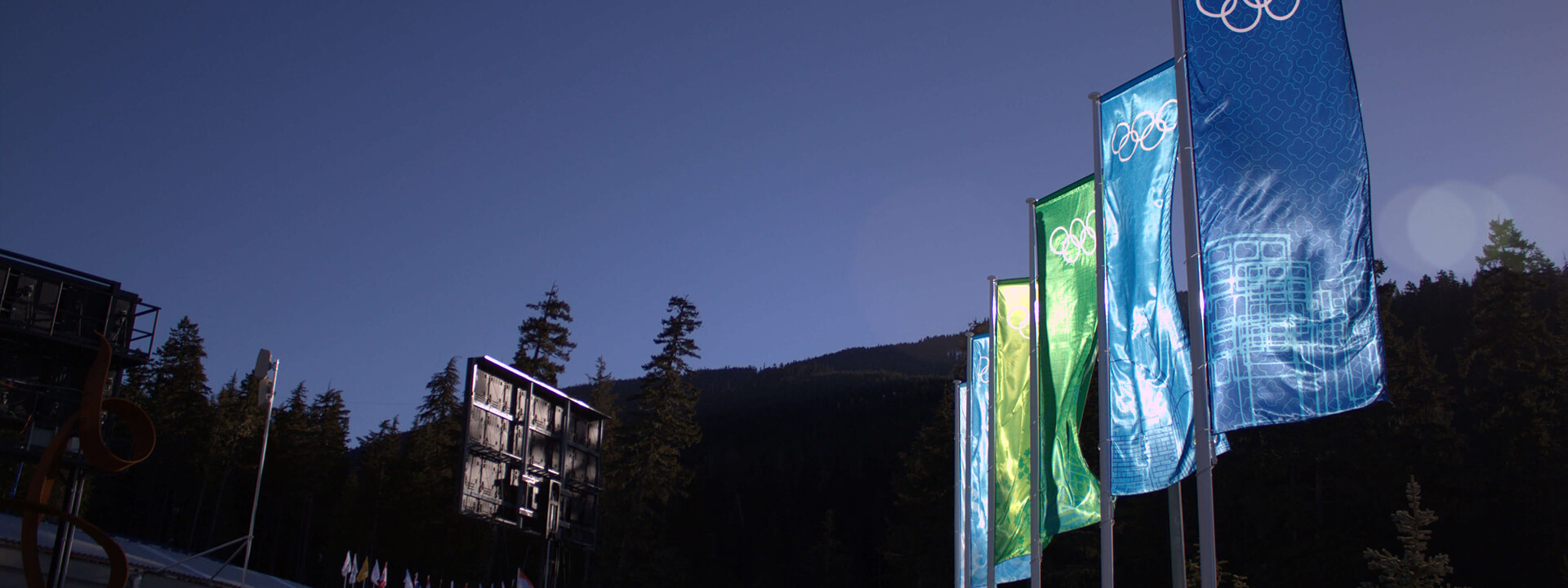 Green and blue Olympic flags wave brightly against a clear blue sky with a dark forested mountainside in the background.