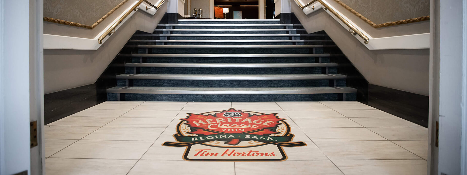 A large floor decal for the NHL Heritage Classic 2019 sponsored by Tim Hortons is prominently displayed on a tiled floor in front of a carpeted staircase.