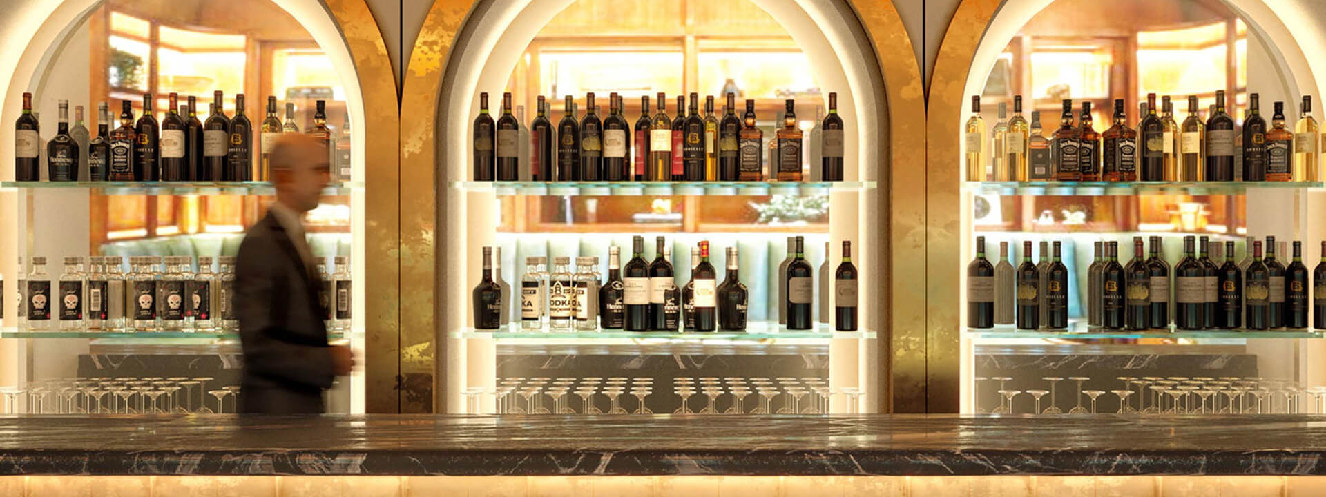 A stylish, well-lit bar features a wide array of alcohol bottles on glass shelves, a dark marble counter lined with glasses, and a blurred person in the background.