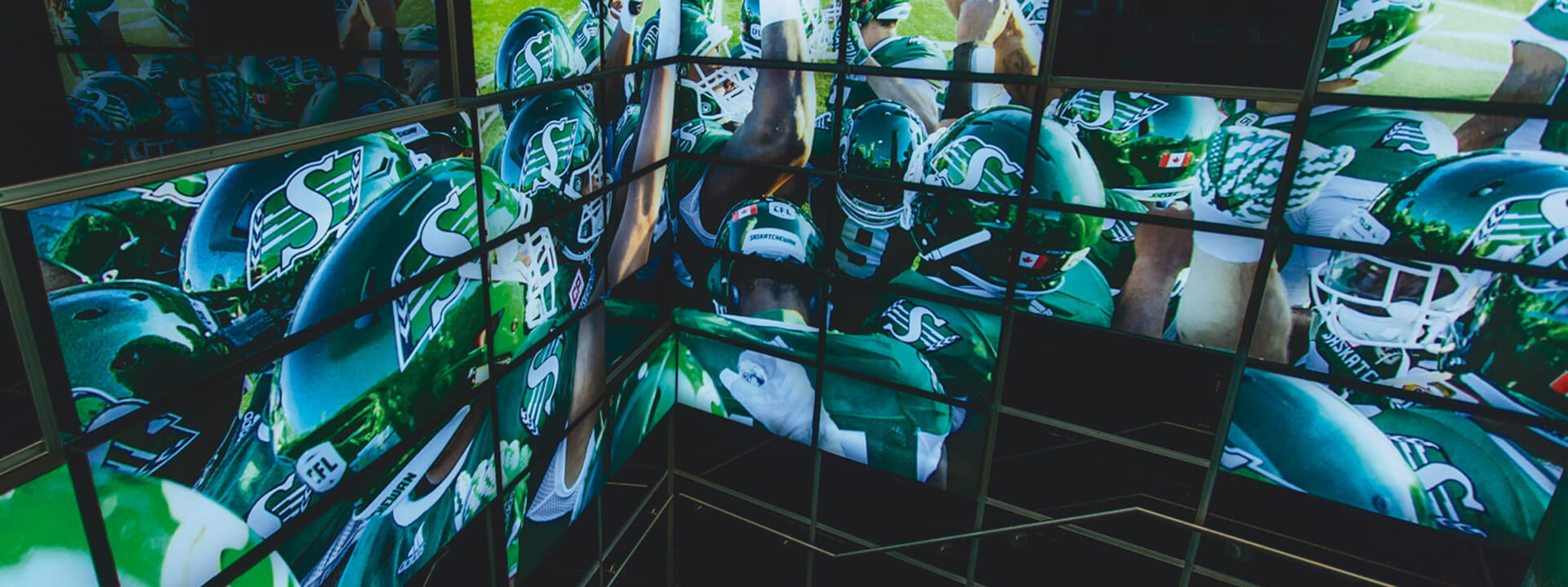 A dynamic corner video wall displays close-up action shots of football players in green Saskatchewan Roughriders uniforms and helmets.