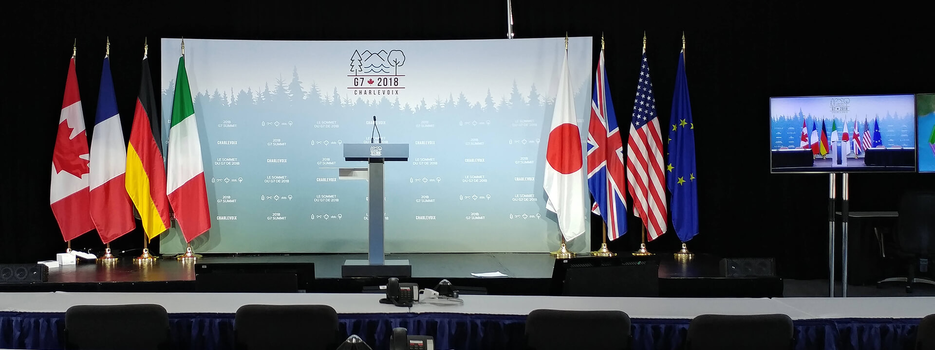 A press conference stage with a podium, flags of G7 nations and the European Union, and a backdrop for the G7 2018 Charlevoix Summit.
