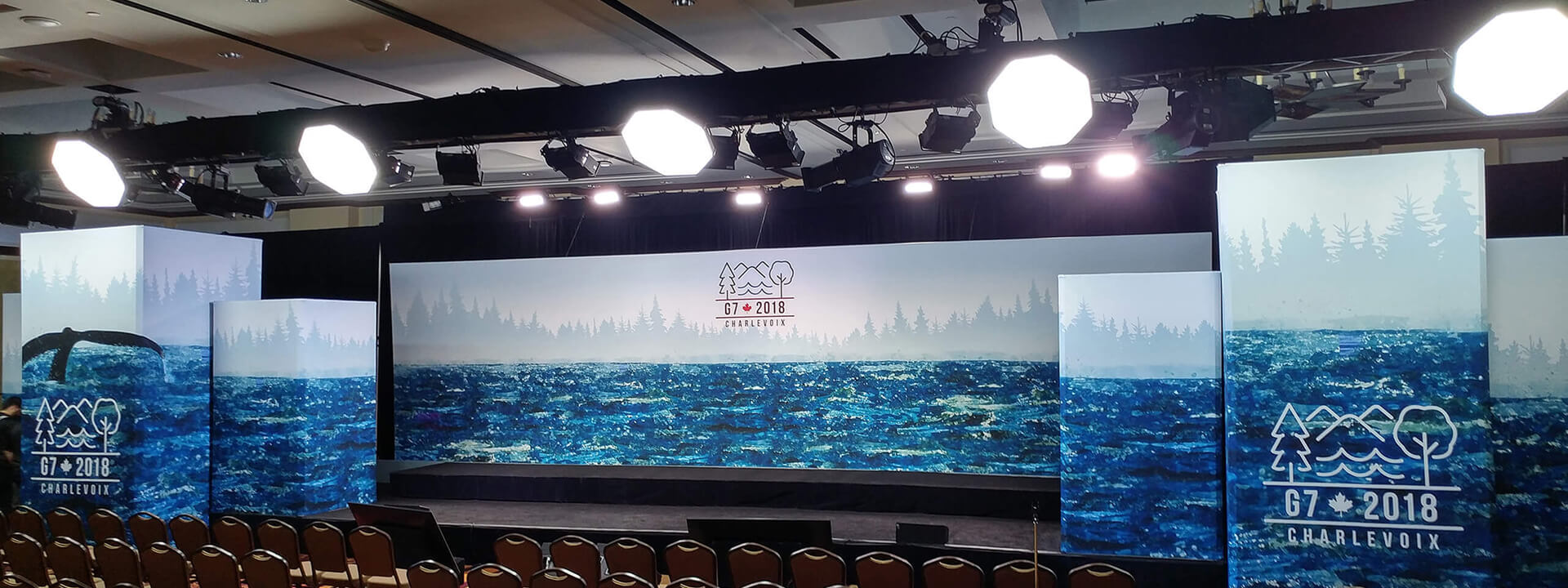 The G7 2018 Charlevoix summit stage features a forest and ocean backdrop with a whale tail and rows of empty chairs.