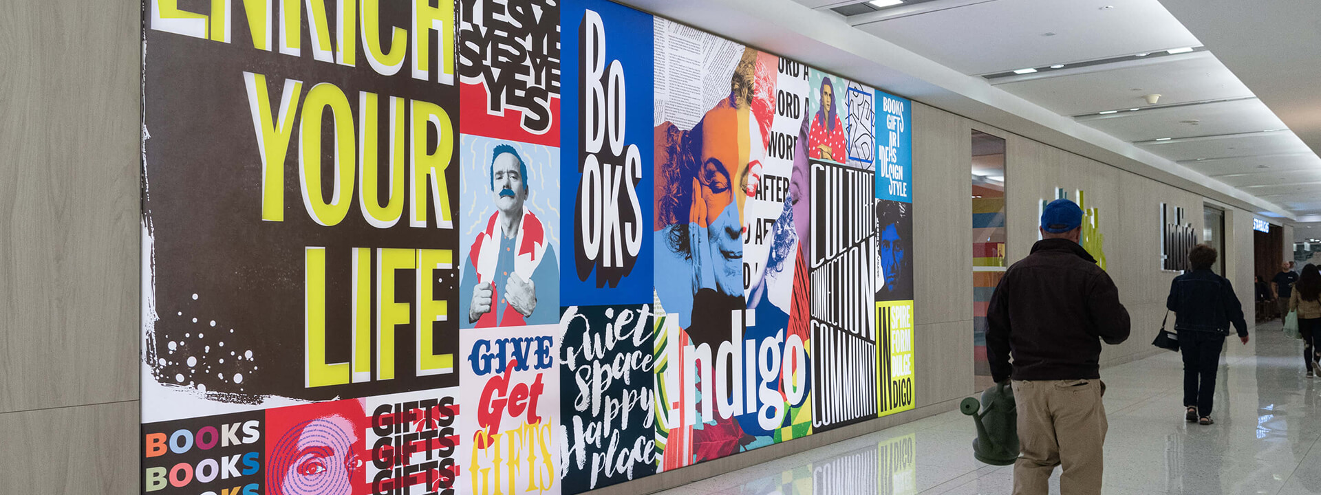 A long, colorful collage mural featuring various posters with text like ""Enrich Your Life,"" ""Books,"" and ""Indigo"" is displayed on a wall in a bright hallway with people walking past.