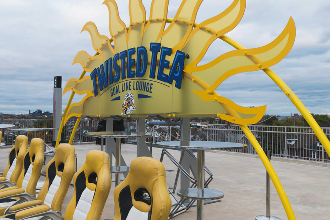 An outdoor lounge area features yellow and black bucket seats, tables, and a large sun-shaped ""Twisted Tea Goal Line Lounge"" sign against a cloudy sky.