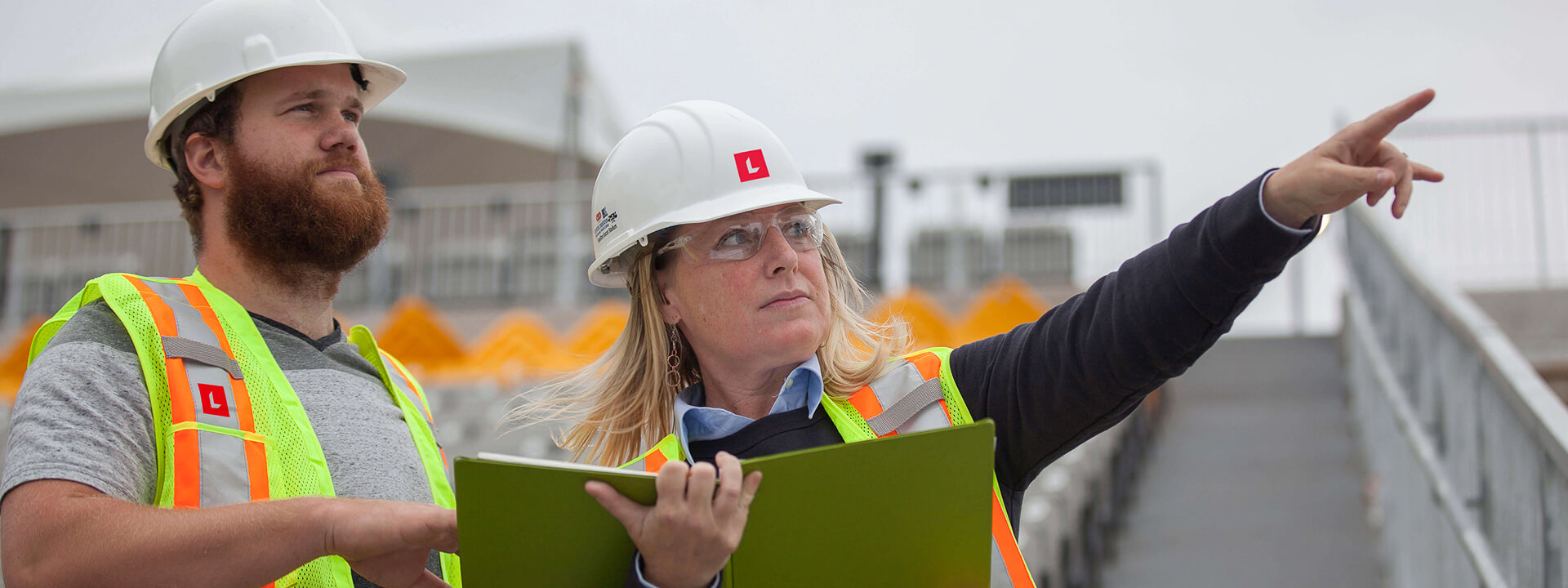 A man and a woman in hard hats and safety vests inspect a worksite, with the woman pointing into the distance and holding a folder.