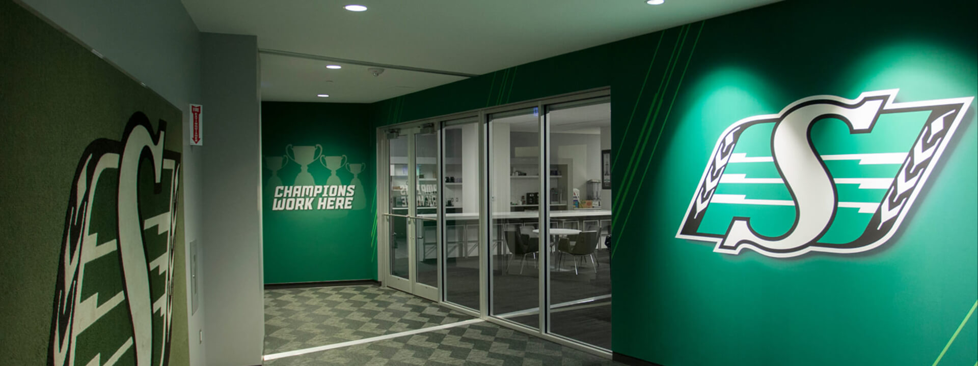 A green and white hallway features the Saskatchewan Roughriders logo, glass office doors, and a wall graphic stating ""CHAMPIONS WORK HERE.""