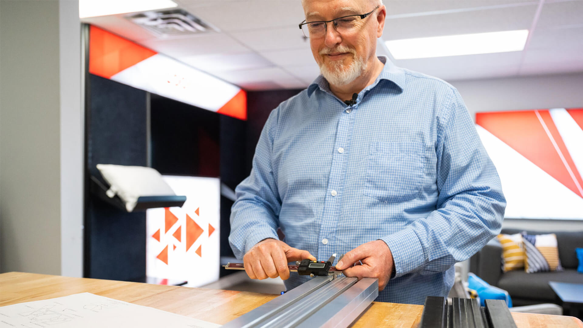 A smiling bearded man uses a digital caliper to measure a metal part on a table.